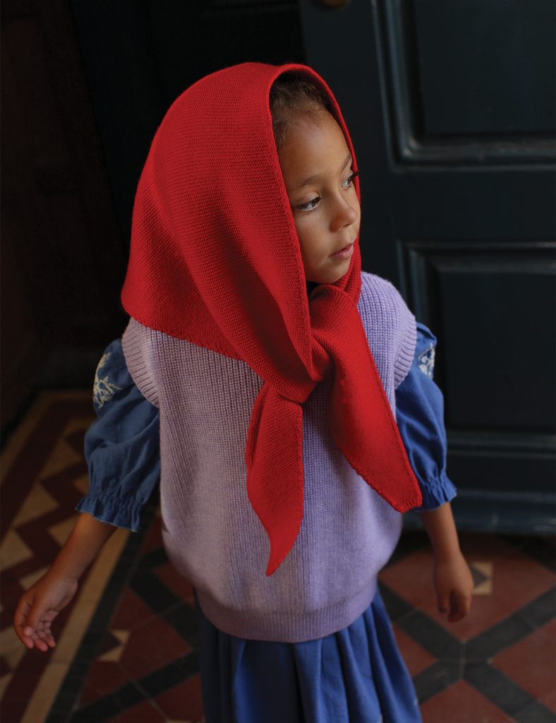 Child wearing a red headscarf and blue dress standing in a room with dark walls and patterned floor.