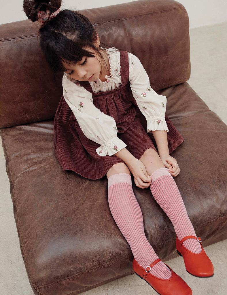 Young girl sitting on a brown couch wearing a burgundy dress, white blouse, pink knee-high socks, and red shoes.