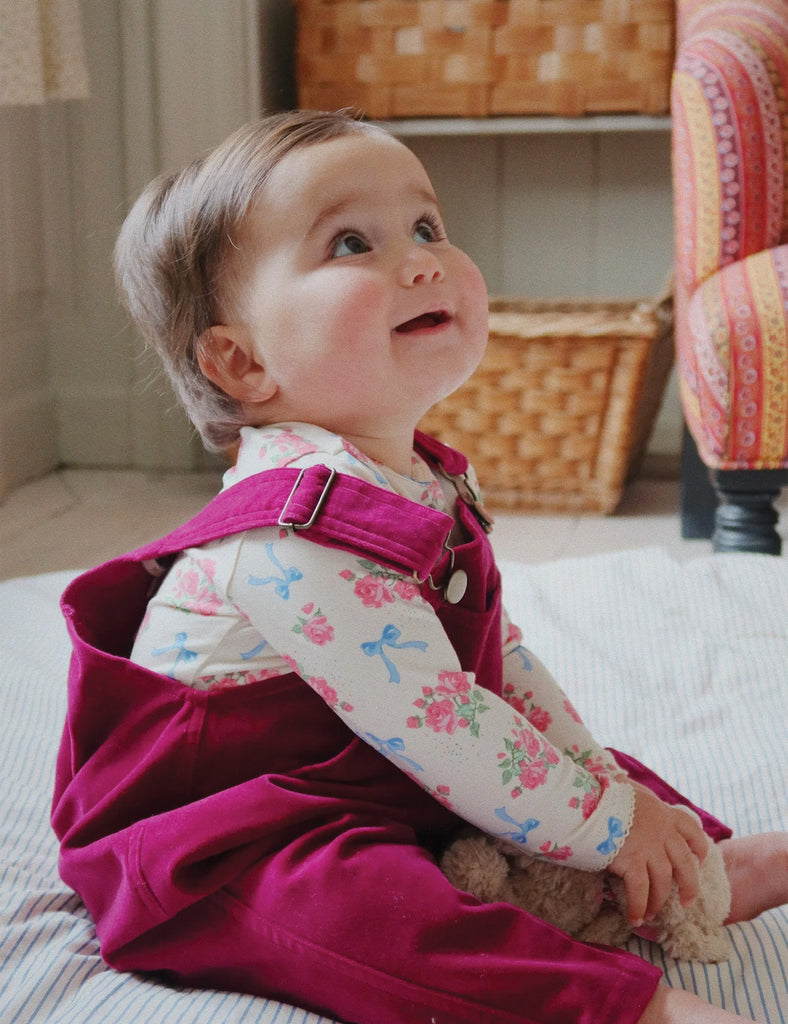 Image of a baby wearing a long sleeve bodysuit with a floral and bow print. Laying down on a bed holding a stuffed puppy.
