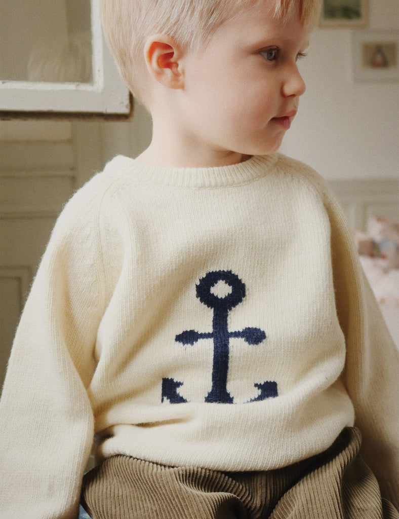 Child wearing a cream sweater with a navy blue anchor design in a home setting.