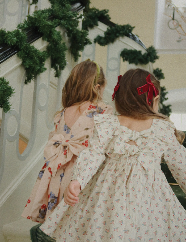 Two young girls in floral dresses with bows standing on a staircase decorated with greenery.