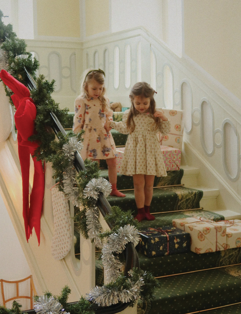 Two young girls in matching dresses standing on a staircase decorated with Christmas decorations and presents.
