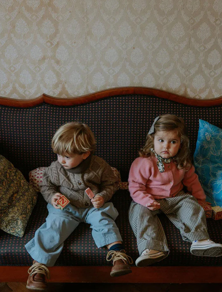 Two children sitting on a couch with patterned wallpaper in the background