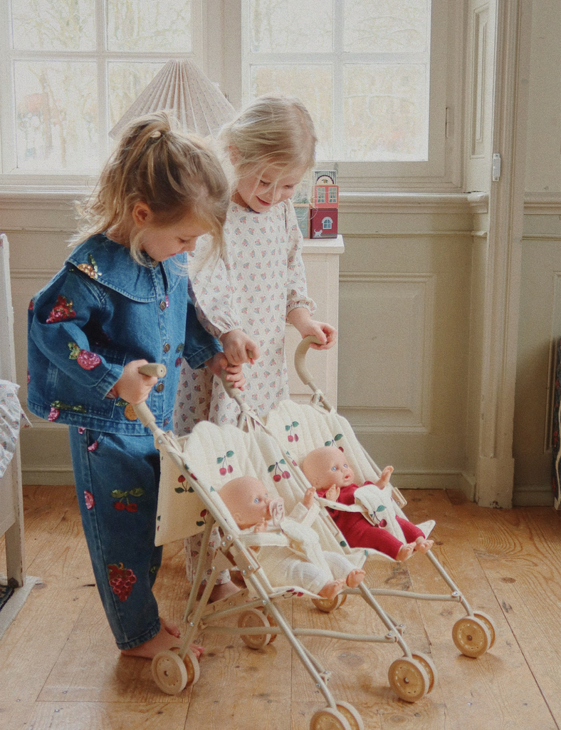 Two young children playing with a doll stroller in a room with large windows.