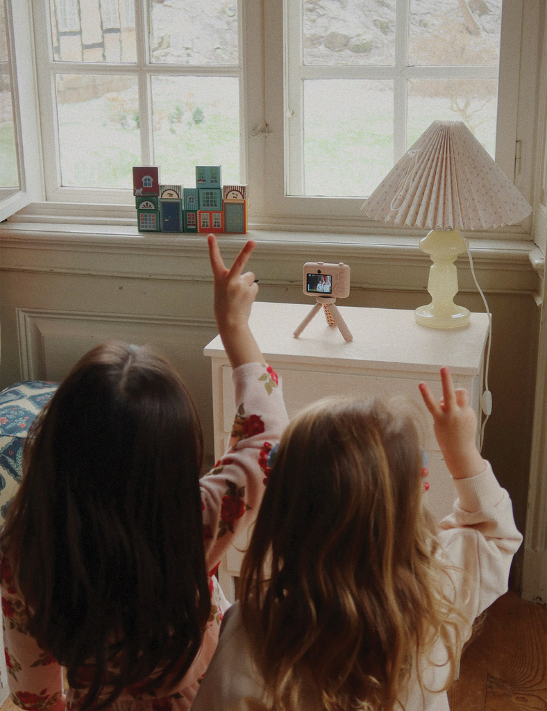 Two children making peace signs in front of a camera on a small table by a window.