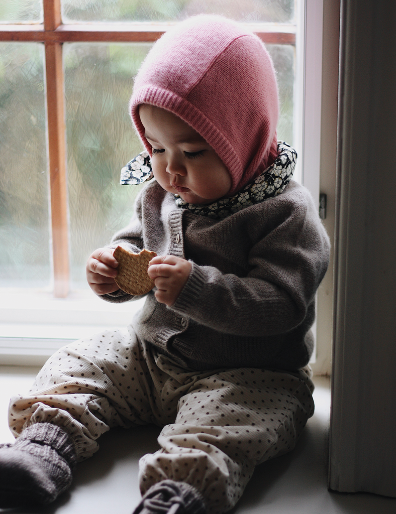 Child wearing a pink knit hat and brown outfit sitting by a window.