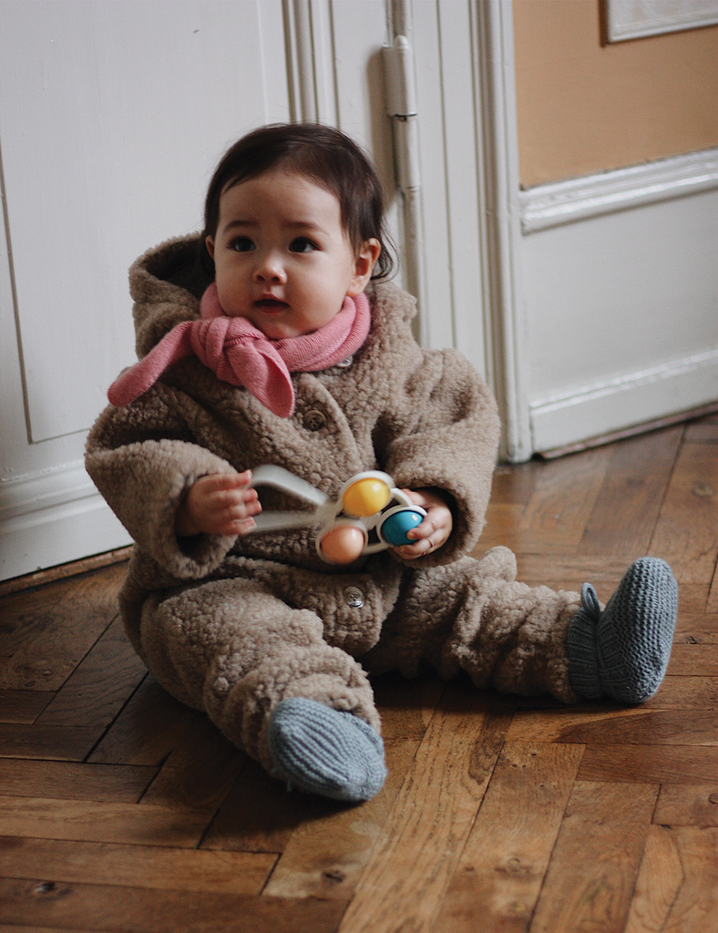 Child in a brown outfit with a pink scarf holding toys on a wooden floor.
