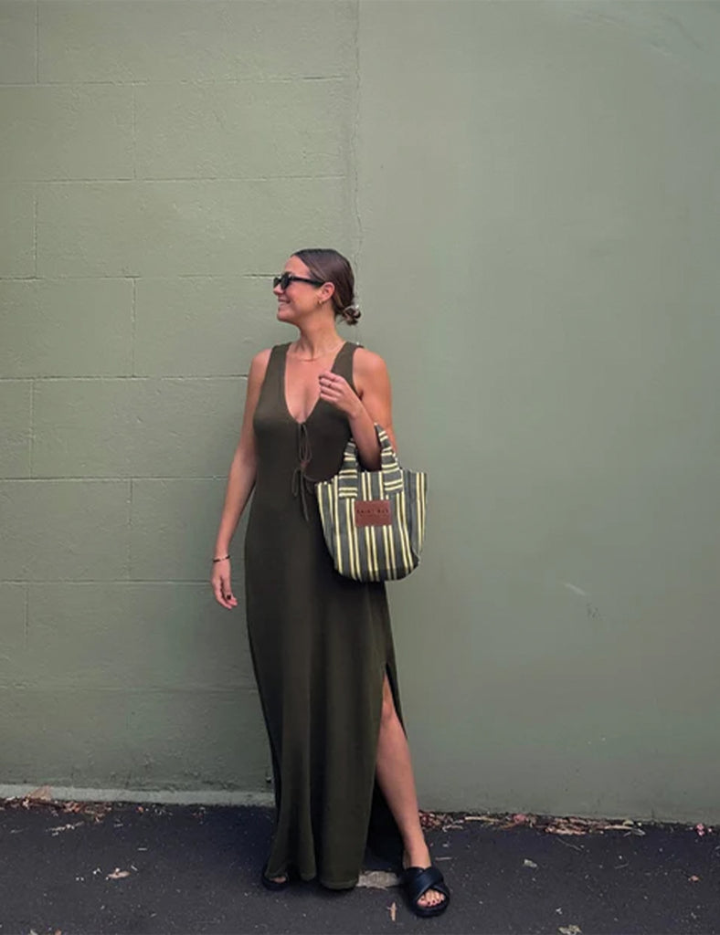 Woman in a green dress holding a striped bag against a green wall.
