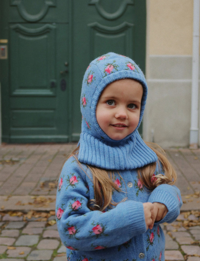 Child wearing a blue knitted balaclava with floral patterns in front of a green door.