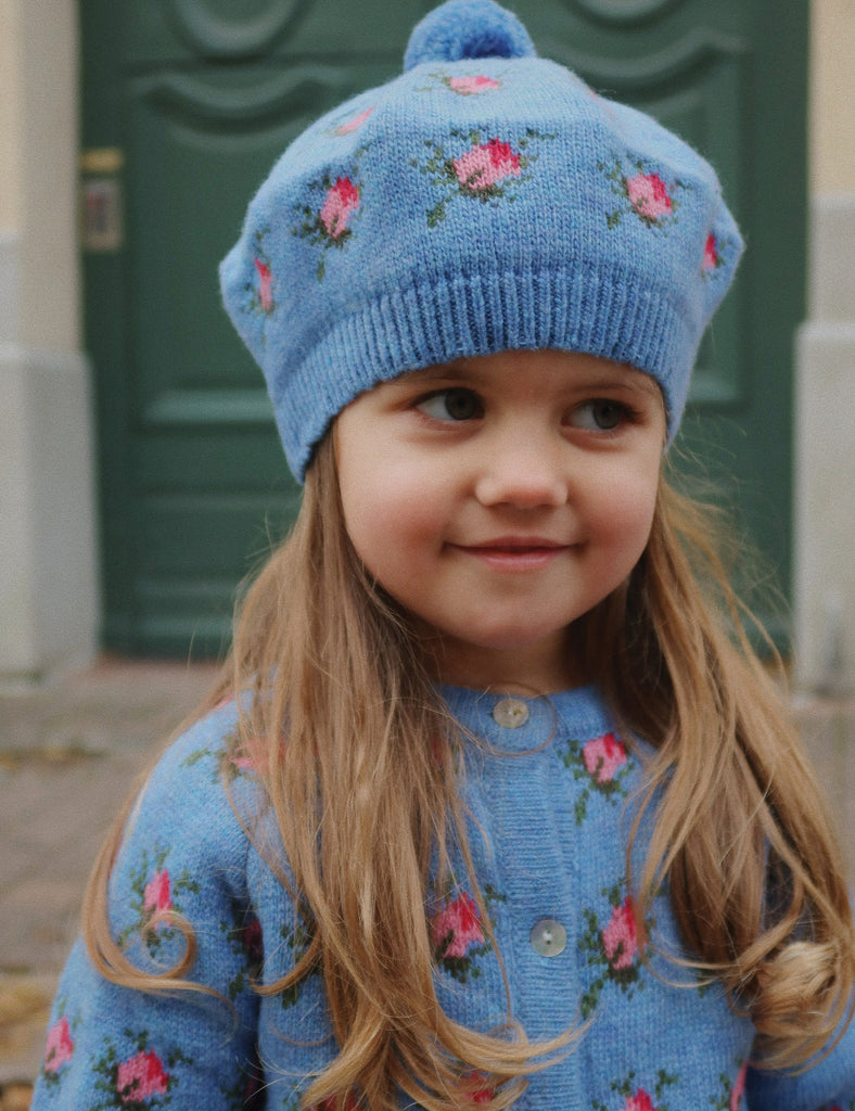 Young girl wearing a blue knitted hat and sweater with floral patterns in front of a green door.