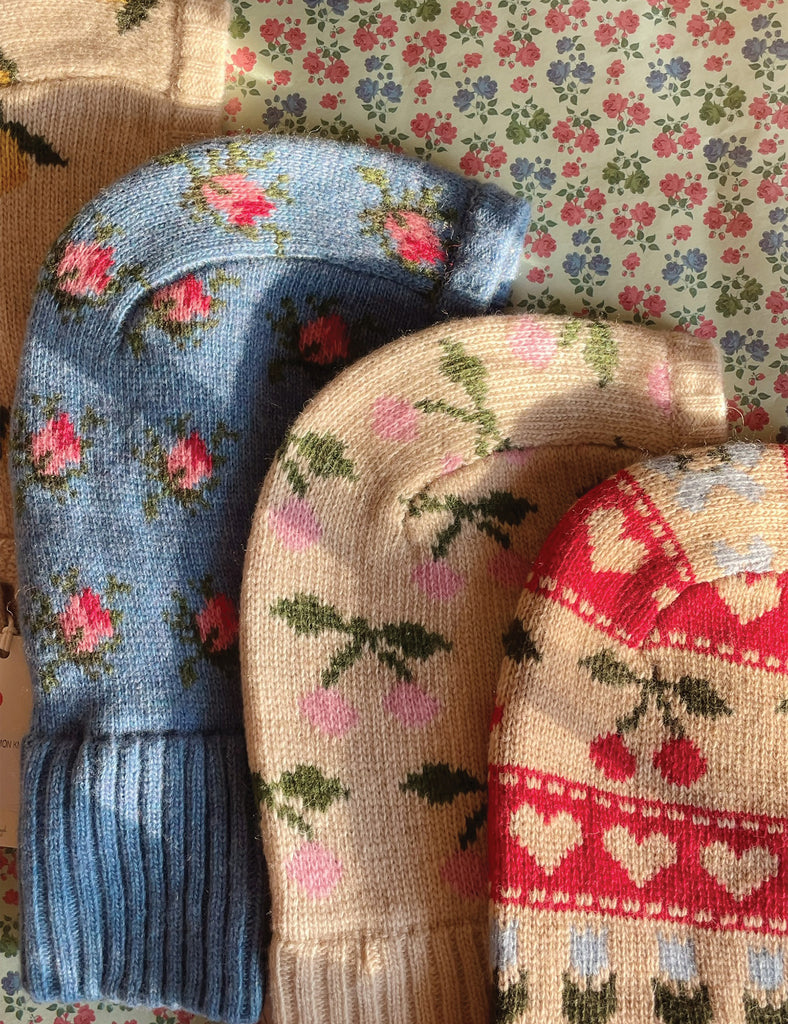 Three knitted balaclavas with floral, cherry and heart patterns on a floral background.