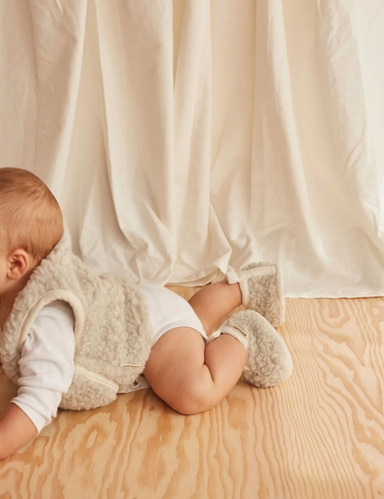 Baby in a white outfit and fluffy socks sitting on a wooden floor with white curtains in the background.