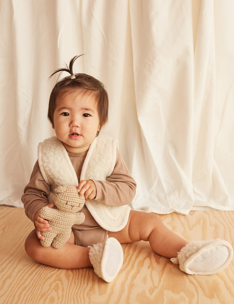 Baby sitting on a wooden floor wearing a brown outfit with a white collar against a beige curtain background.