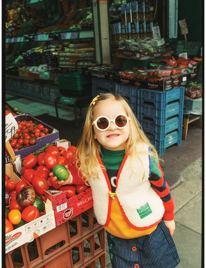 Young girl in a colorful outfit with sunglasses standing in front of a produce stand.