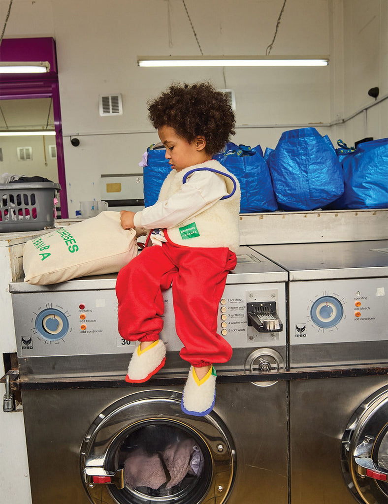 Child sitting on a washing machine in a laundromat with bags of laundry.