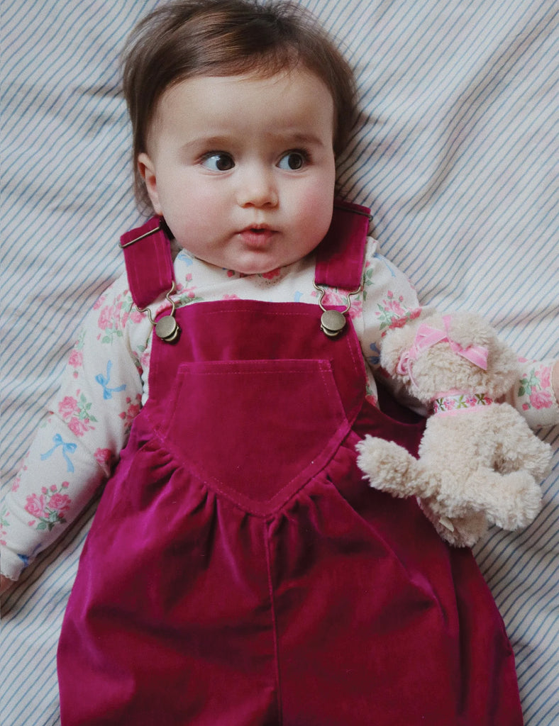 Image of a baby wearing a long sleeve bodysuit with a floral and bow print. Laying down on a bed holding a stuffed puppy.