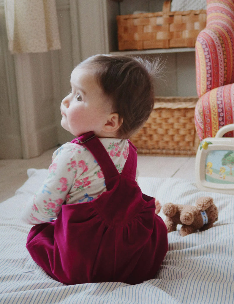 Image of a baby wearing a long sleeve bodysuit with a floral and bow print. Laying down on a bed holding a stuffed puppy.