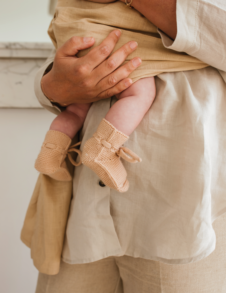 Person holding a baby wearing beige knitted booties with a neutral background