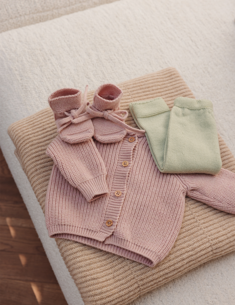 Image of pink booties and baby cardigan laying on top of a knitted blanket.
