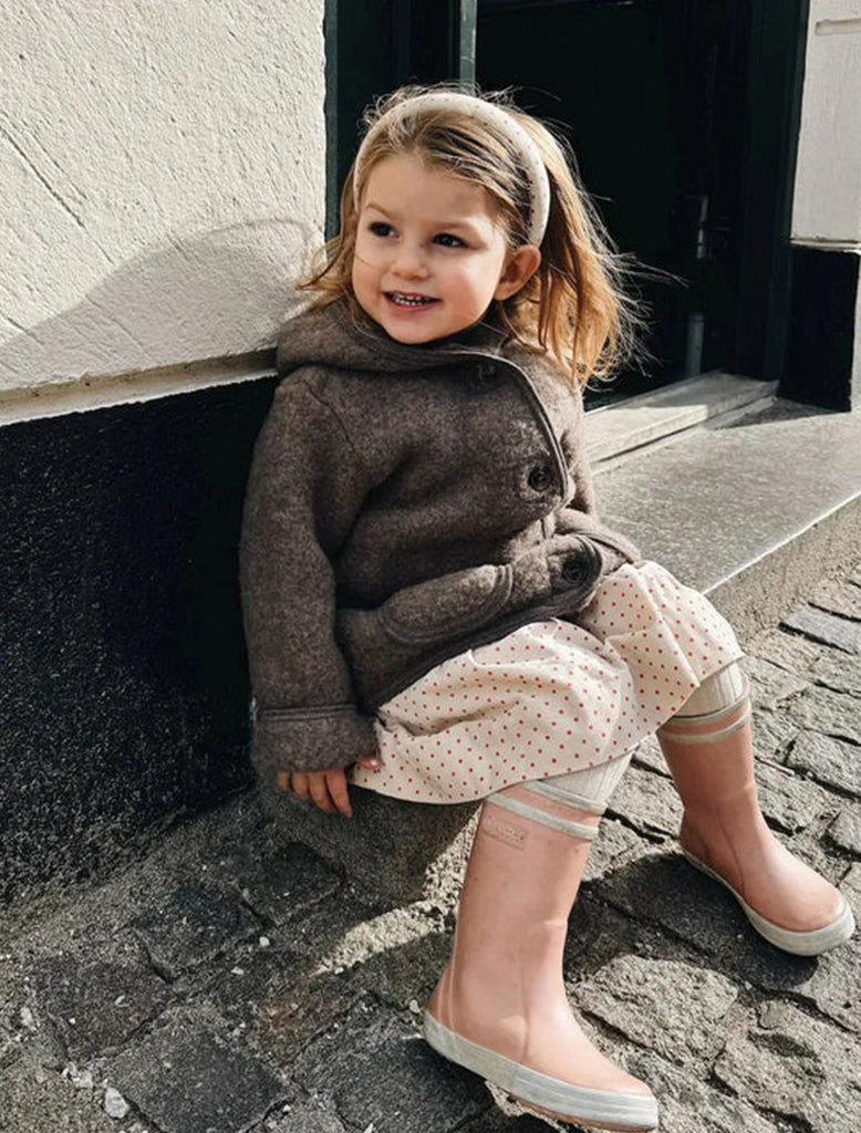 Young girl sitting on a stone pavement wearing a brown sweater and pink boots.
