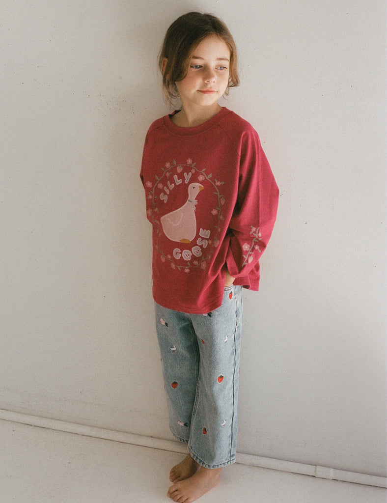 Child wearing a red sweatshirt with a graphic design and light blue jeans against a white wall.