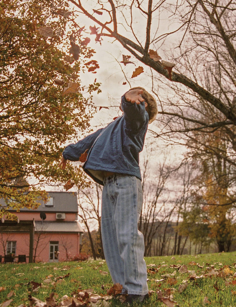Person in a blue sweater and jeans standing in a park with trees and a house in the background.