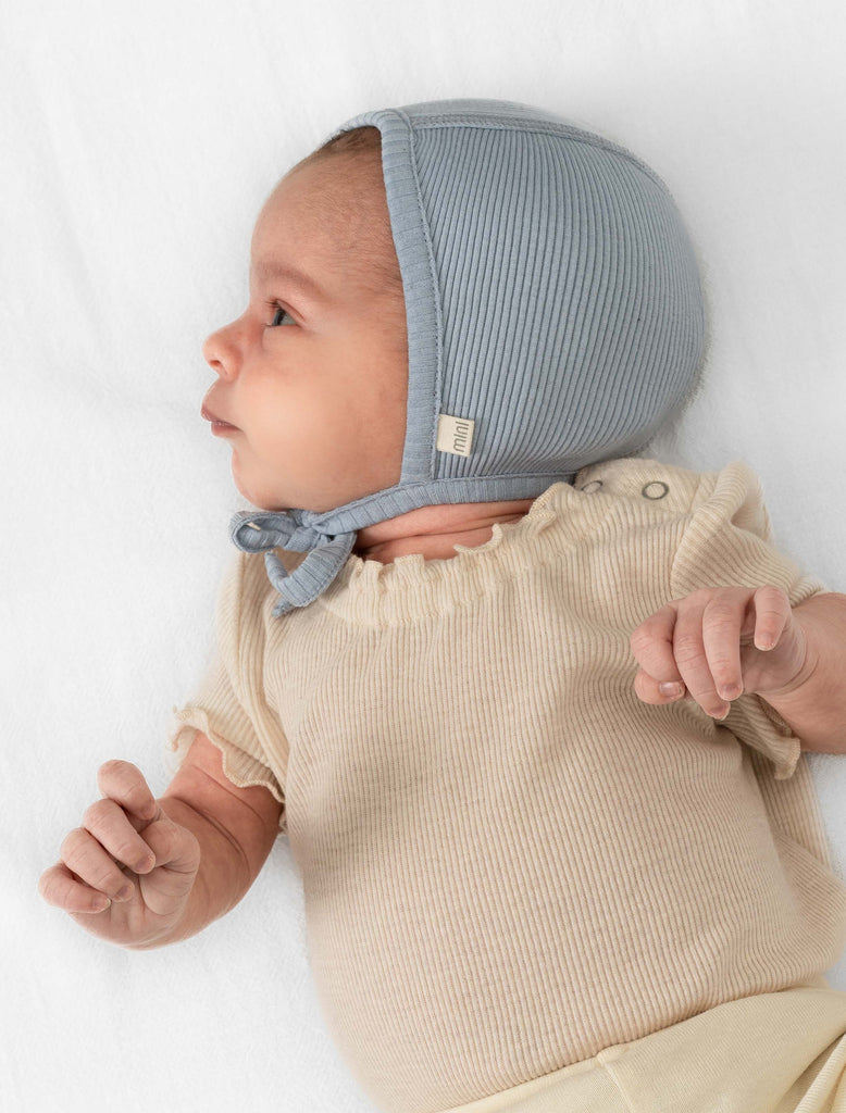 This image shows a newborn baby wearing the Bloom Silk Knit Bonnet in Clearwater Blue. The baby is laying down wearing an ivory onesie and the bonnet.