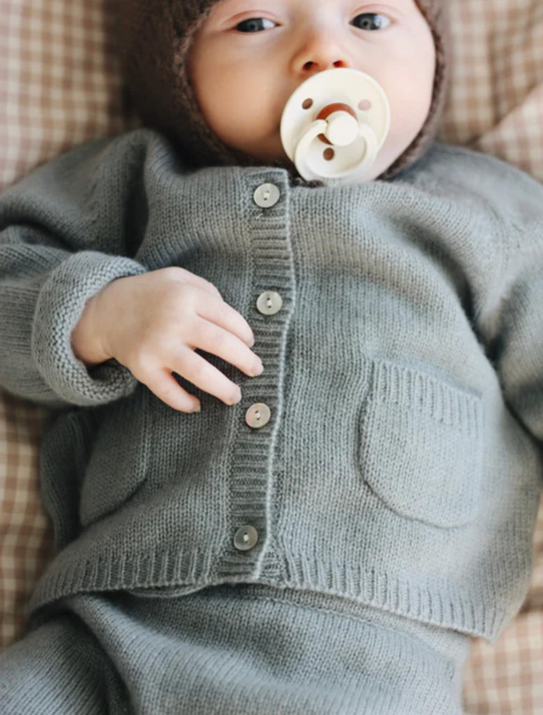 Baby wearing a gray knitted cardigan with a pacifier in mouth, lying on a checkered surface.