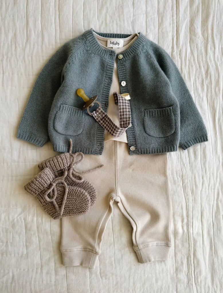Children's outfit with a green knitted cardigan, beige pants, and brown knitted booties on a light wooden surface.
