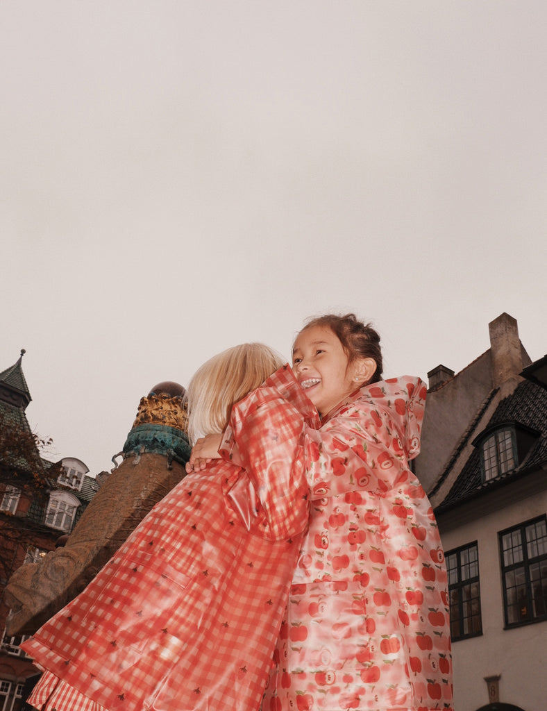 Two children in red checkered raincoats standing in front of a building with a light-colored wall and dark roof.