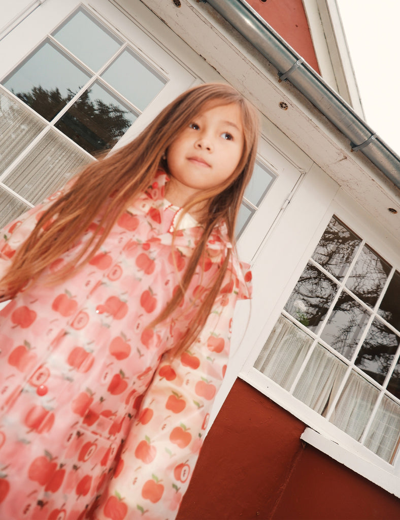 Young girl in a rain coat with red patterns standing in front of a house.