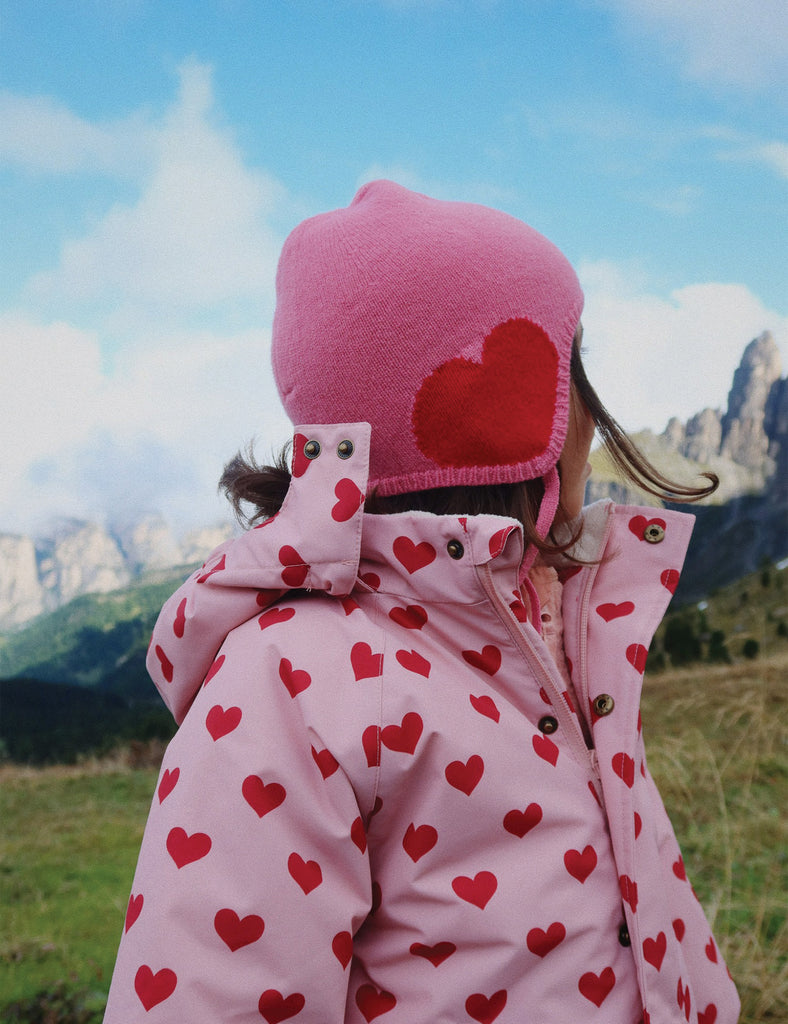 Person wearing a pink jacket with red heart patterns and a matching pink hat in a mountainous landscape.