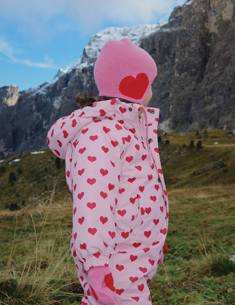 Person wearing a pink snowsuit with red hearts and a matching hat, standing in a mountainous landscape.