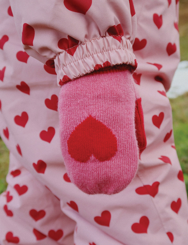 Pink mitten with red heart design worn by a child in a pink outfit with red heart pattern.