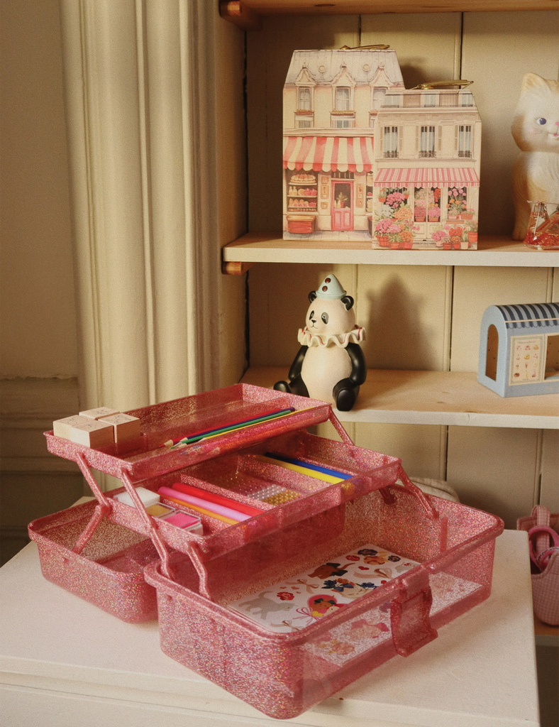 Pink toy storage box with stationery items on a table, shelves with decorative items in the background.