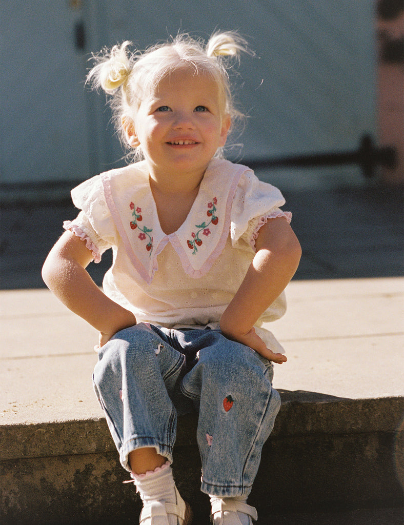 Young girl sitting on a ledge wearing a white blouse with embroidery and blue jeans.