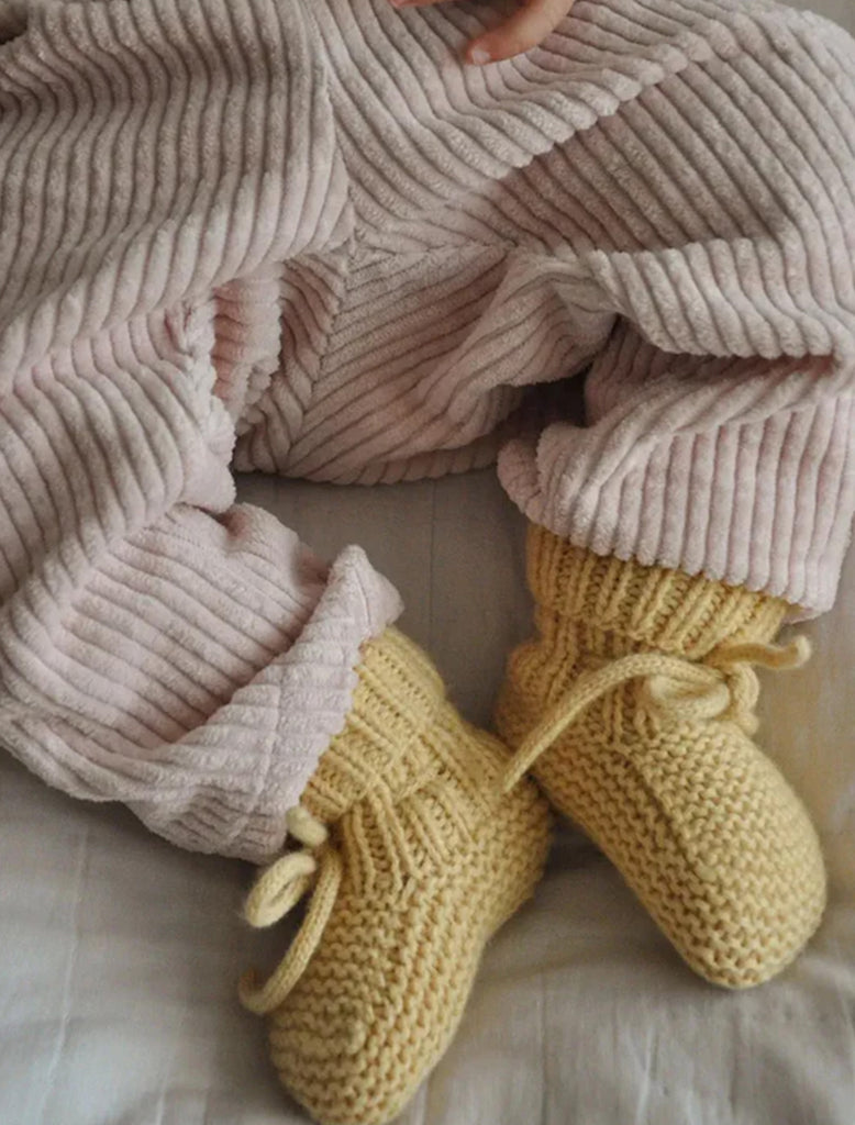 Person wearing beige ribbed pants with yellow knitted baby shoes on a light background