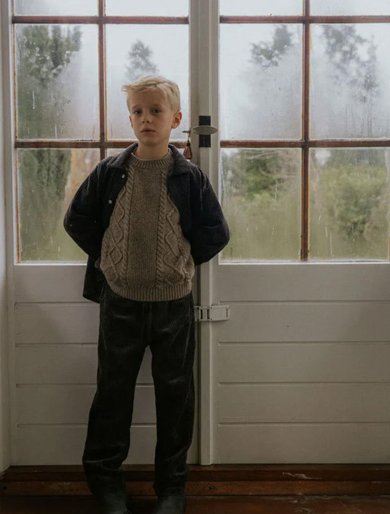 Child wearing a sweater and jacket standing in front of a glass door with a blurred outdoor view.