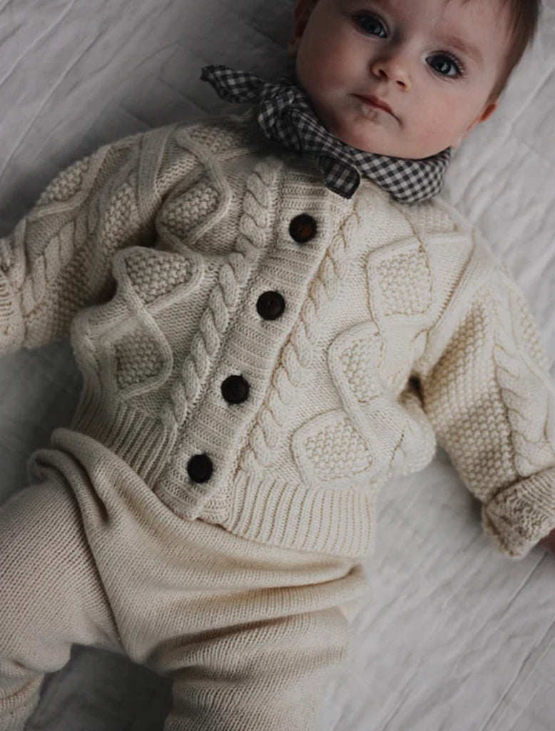 Baby wearing a cream knitted cardigan with buttons on a light wooden floor.