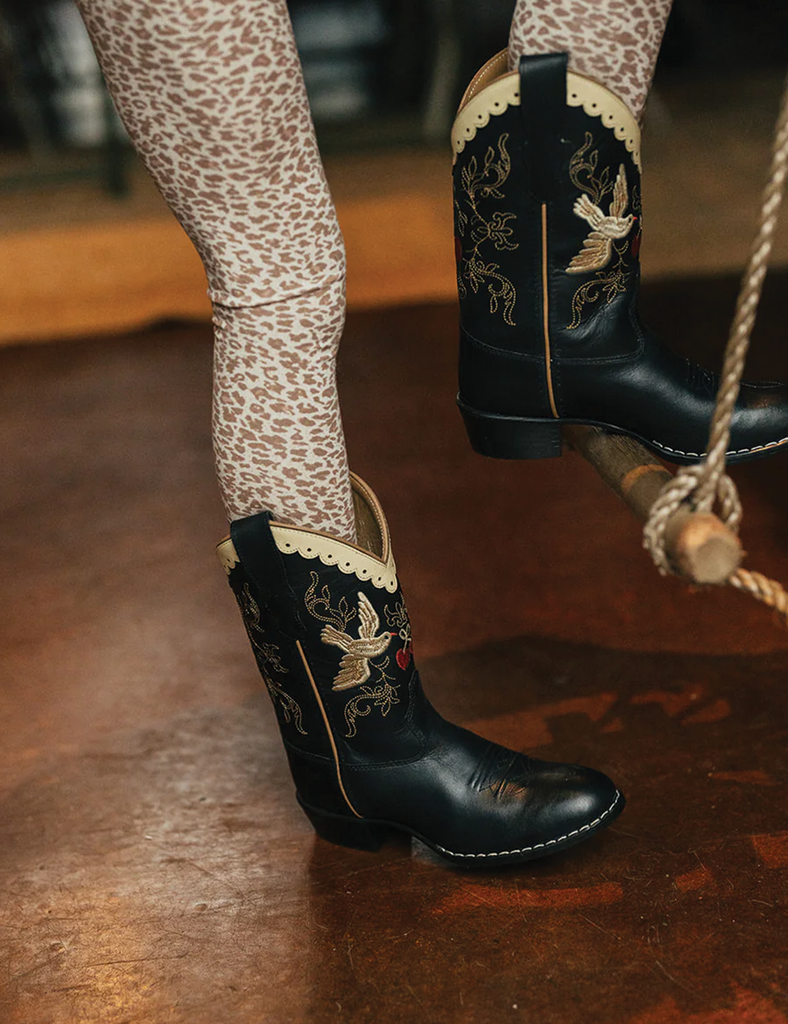 Black embroidered cowboy boots with patterned leggings on a wooden floor.