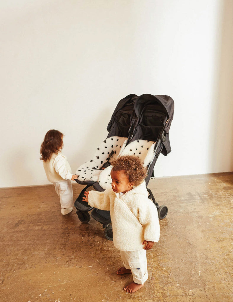 Two children in white outfits with a black stroller on a plain floor.