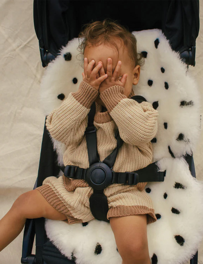 Child in a beige knit outfit sitting in a stroller with a polka dot pattern.