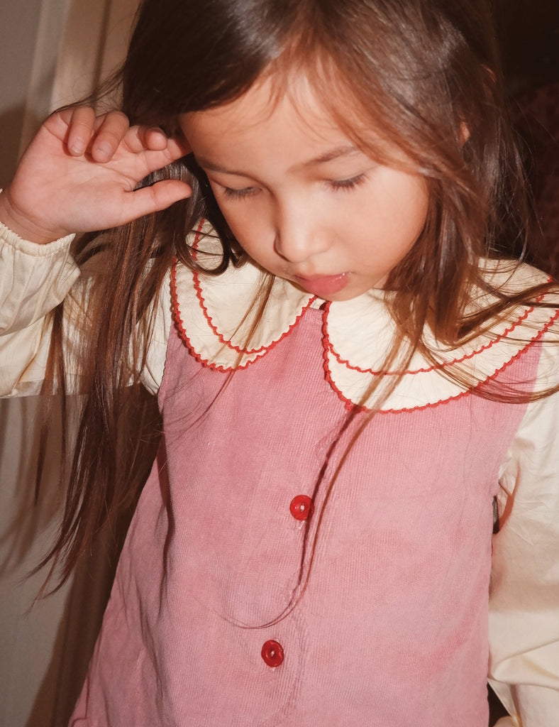 Young girl with long hair wearing a pink dress with red buttons and a white lace collar.