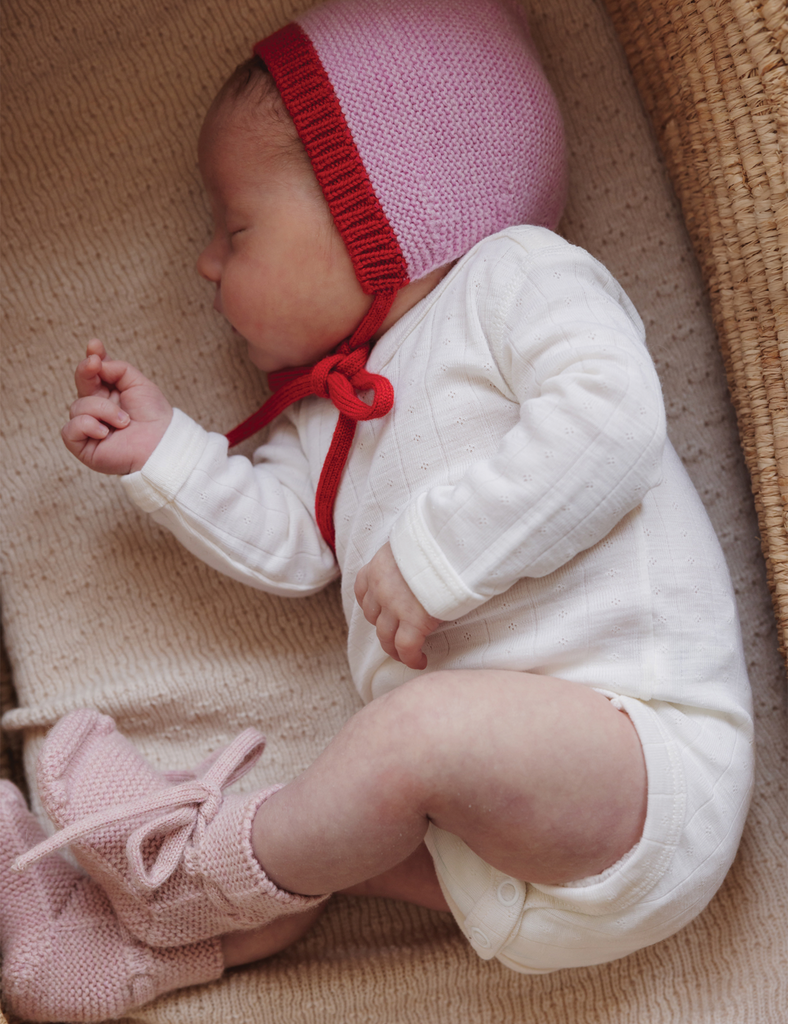 Baby wearing a pink knitted hat with a red band and matching shoes, lying on a textured surface.