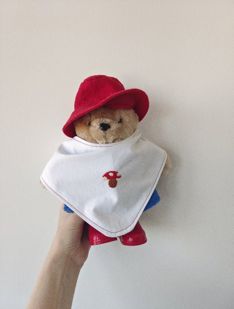 Teddy bear wearing a red hat and bib with a mushroom design, held against a plain background.