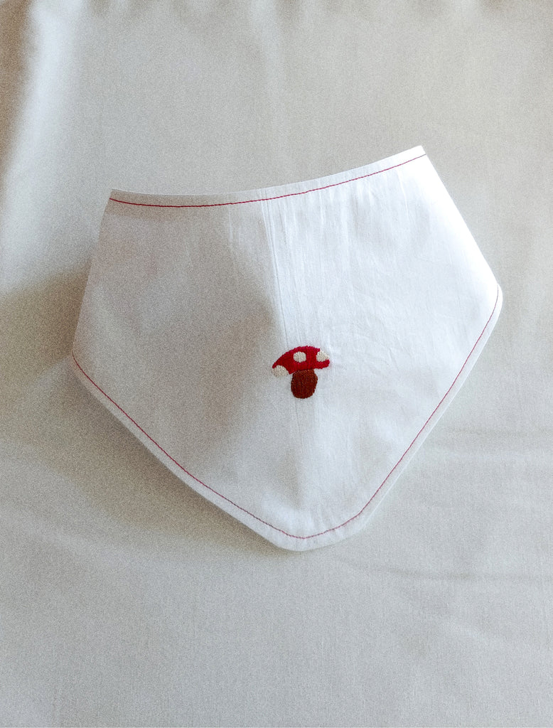 White baby bib with red trim and embroidered mushroom design on a light background