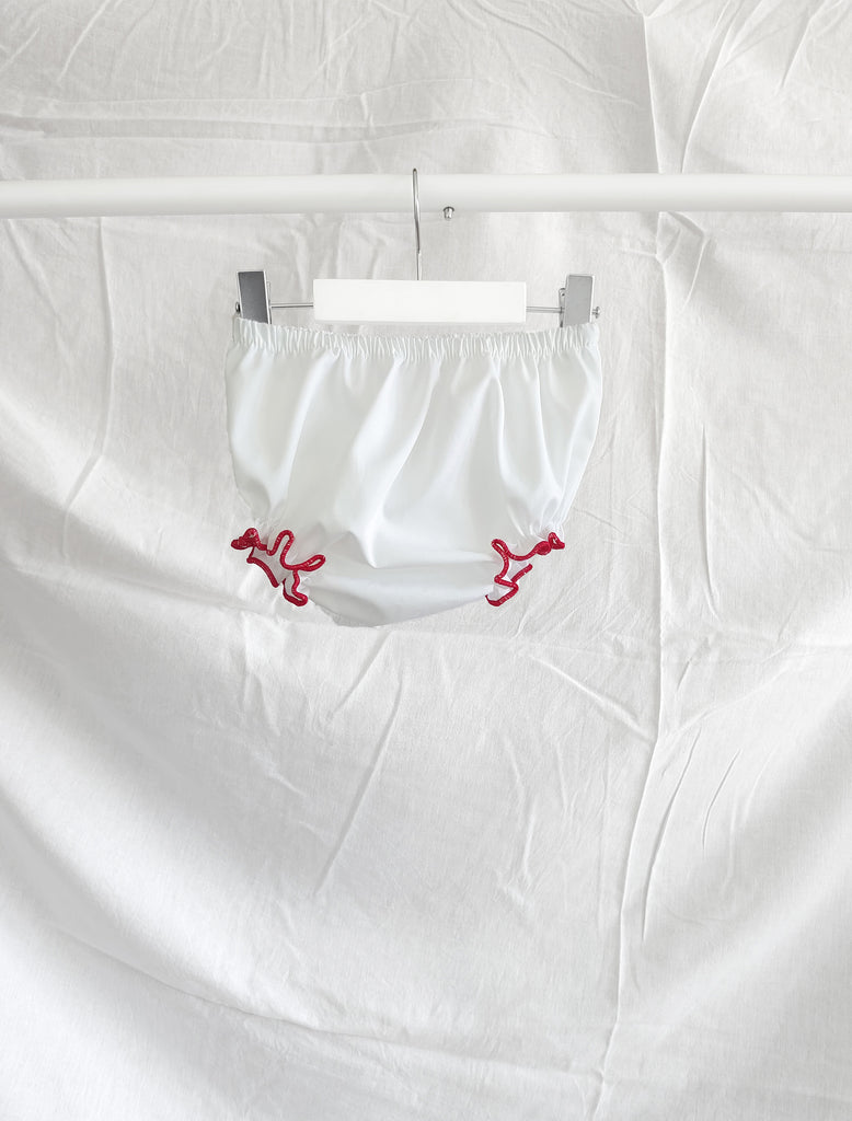 White baby bloomers with contrast red ruffle stitching hanging on a white hanger in front of a white background.