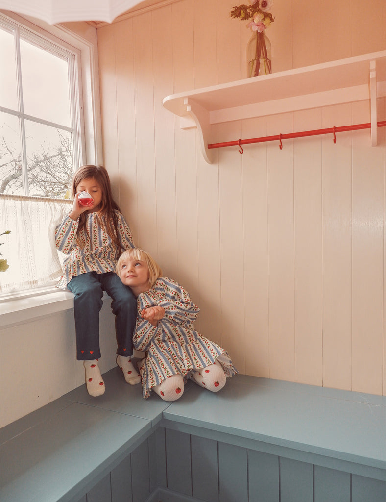 Two children sitting on a window sill in a room with light-colored walls and a red hook.