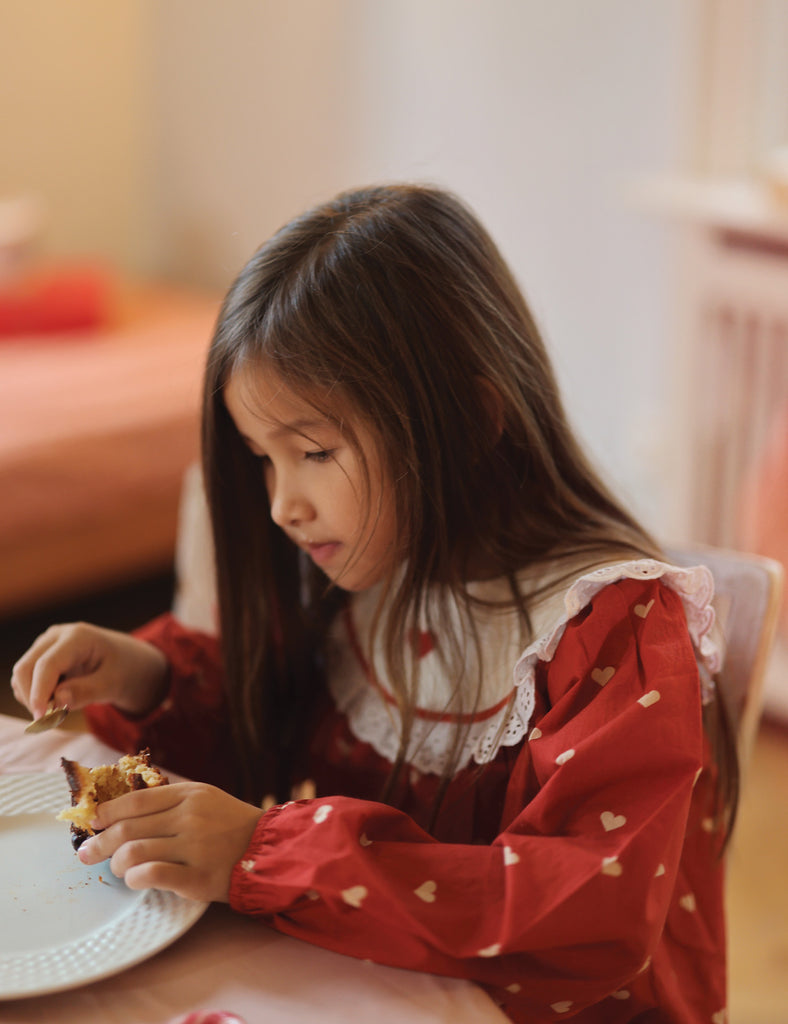 Young girl in a red dress with heart patterns eating at a table.