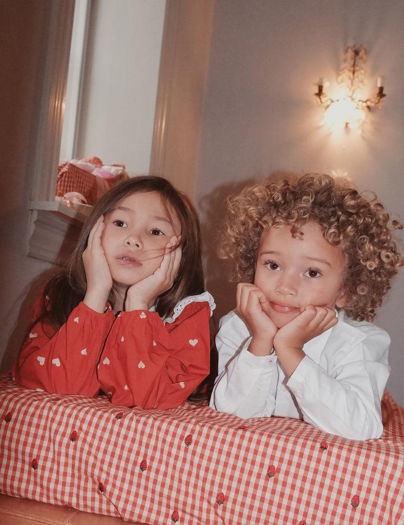Two children sitting on a bed with red and white checkered bedding, one in a red outfit and the other in a white outfit.